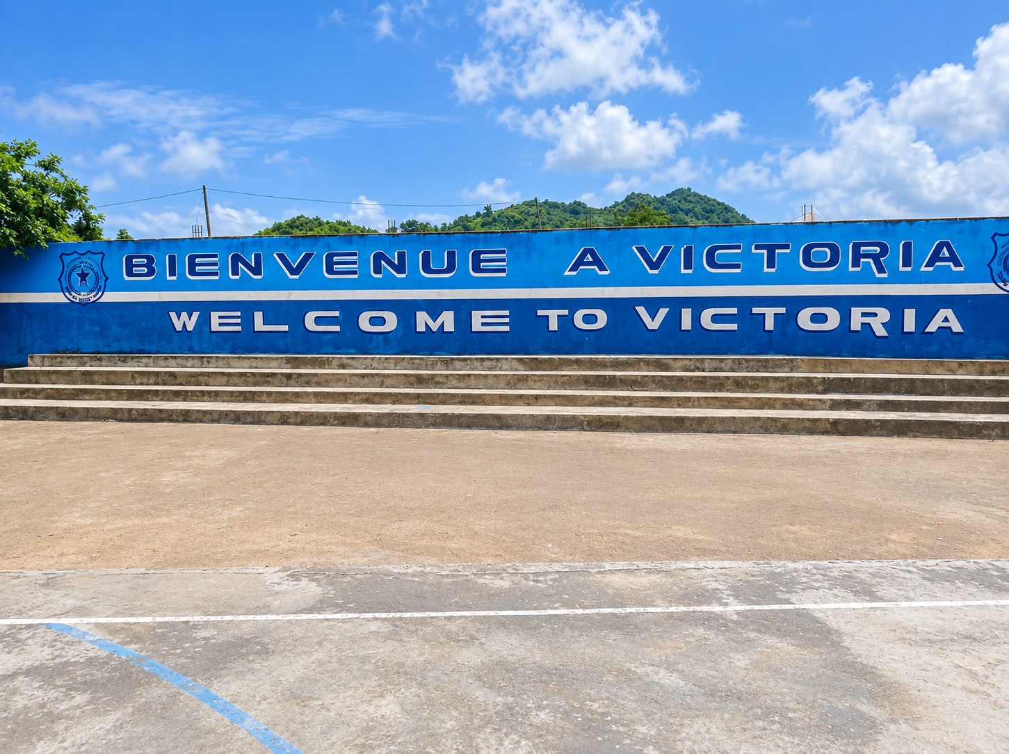 Mur d'enceinte peint en bleu portant « Bienvenue à Victoria / Welcome to Victoria », gradins et esplanade, colline boisée en arrière-plan.
